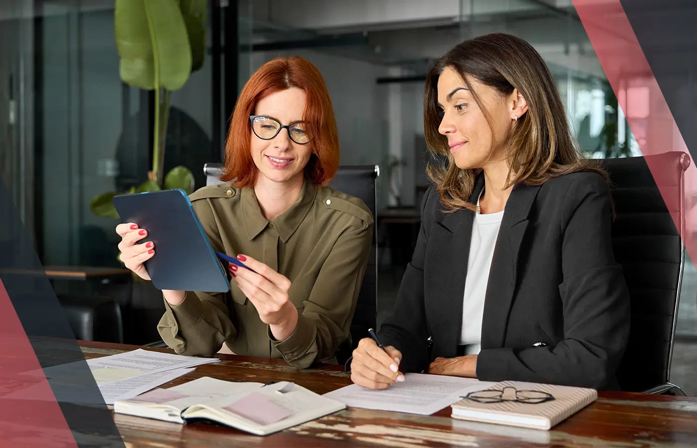 Two professionals in an office reviewing information on a tablet at a desk with papers and notebooks during a business discussion.