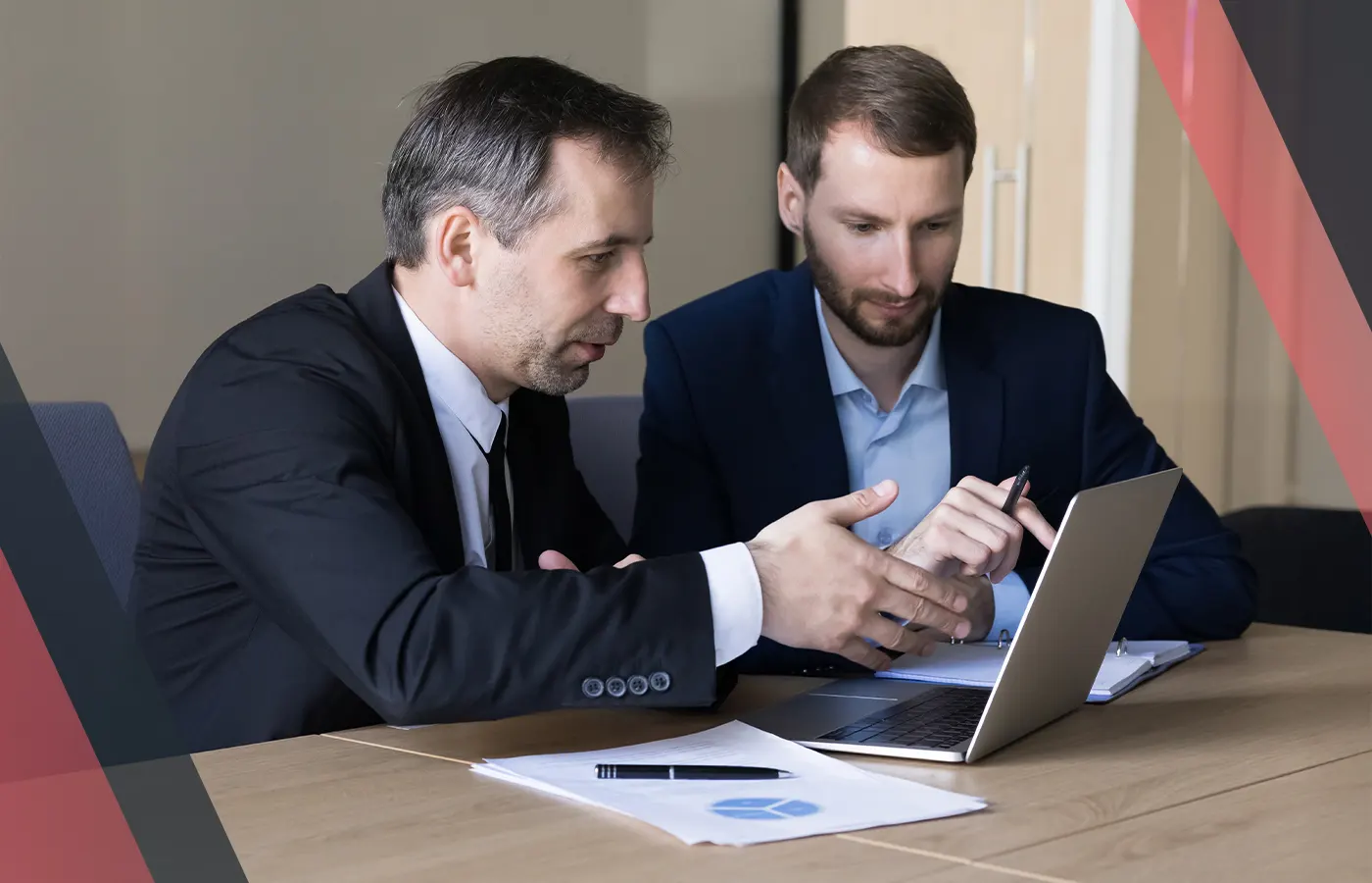 Two professionals in suits discussing at a laptop during a meeting, with paperwork and a notebook on the table.