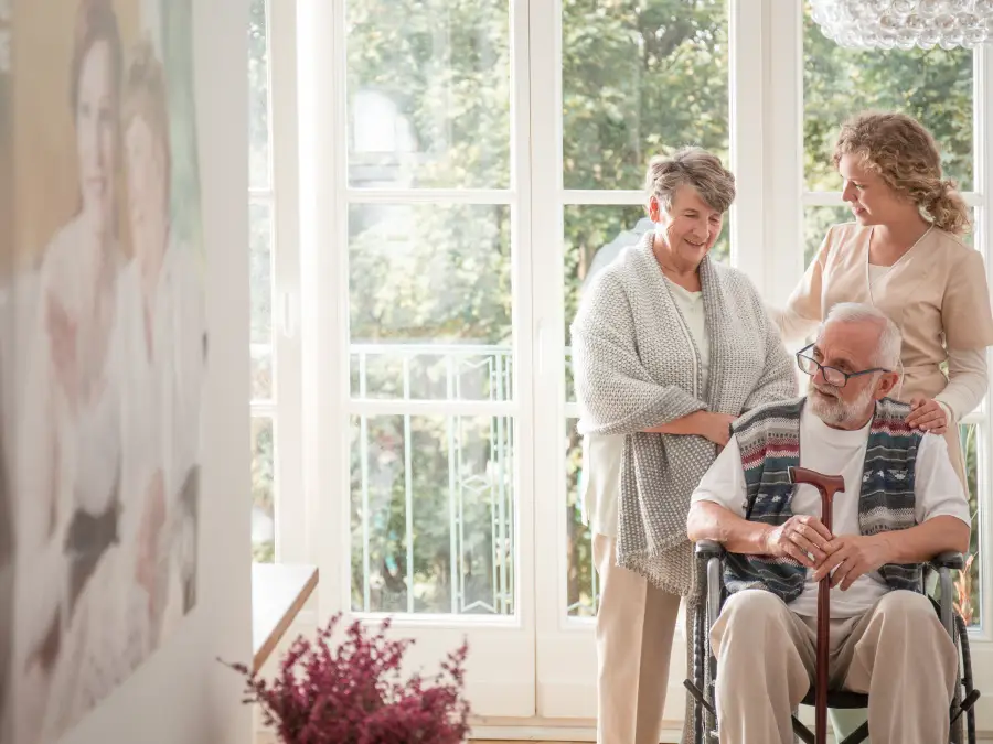 A Senior man with a walking stick sits in a wheelchair while a caregiver and an elderly woman stand behind him outdoors.
