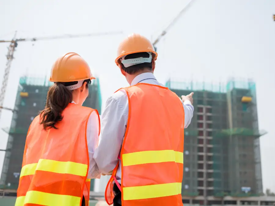 Rear view of two business partners in hard hats looking at a large building under construction at a sunny site.