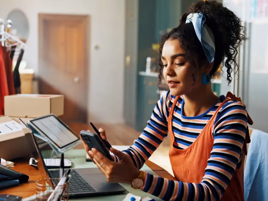 Woman working on multiple gadgets while processing online orders, with the products in the background