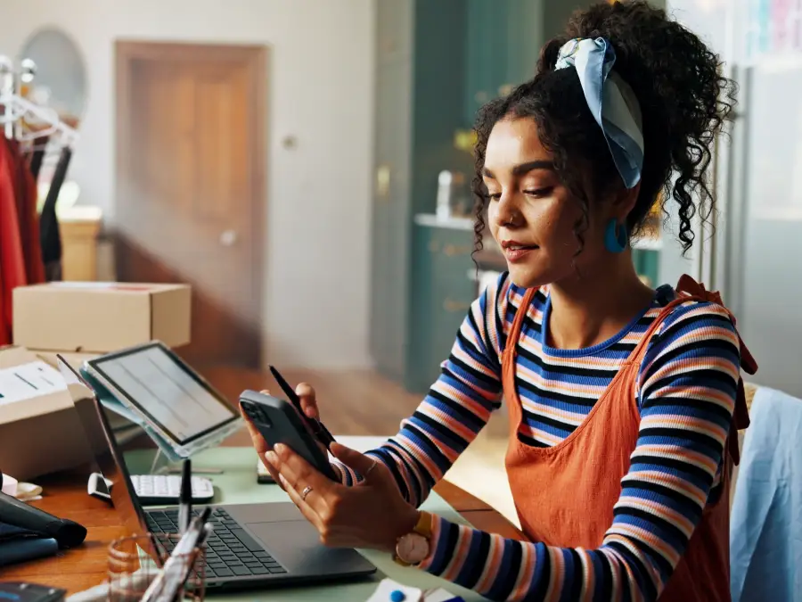 Woman working on multiple gadgets while processing online orders, with the products in the background