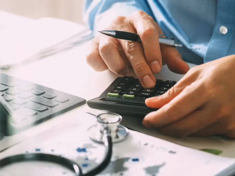 A doctor uses a calculator and laptop next to a stethoscope, focusing on healthcare costs and medical fees.