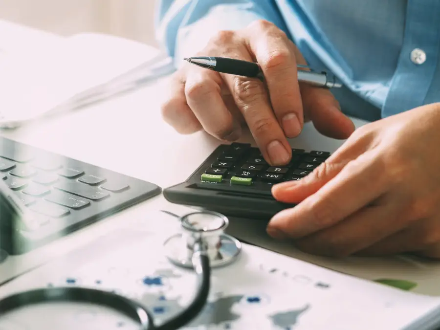 A doctor uses a calculator and laptop next to a stethoscope, focusing on healthcare costs and medical fees.