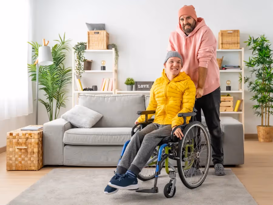 A portrait of a smiling caregiver standing behind a senior man in a wheelchair, both looking at the camera outdoors.