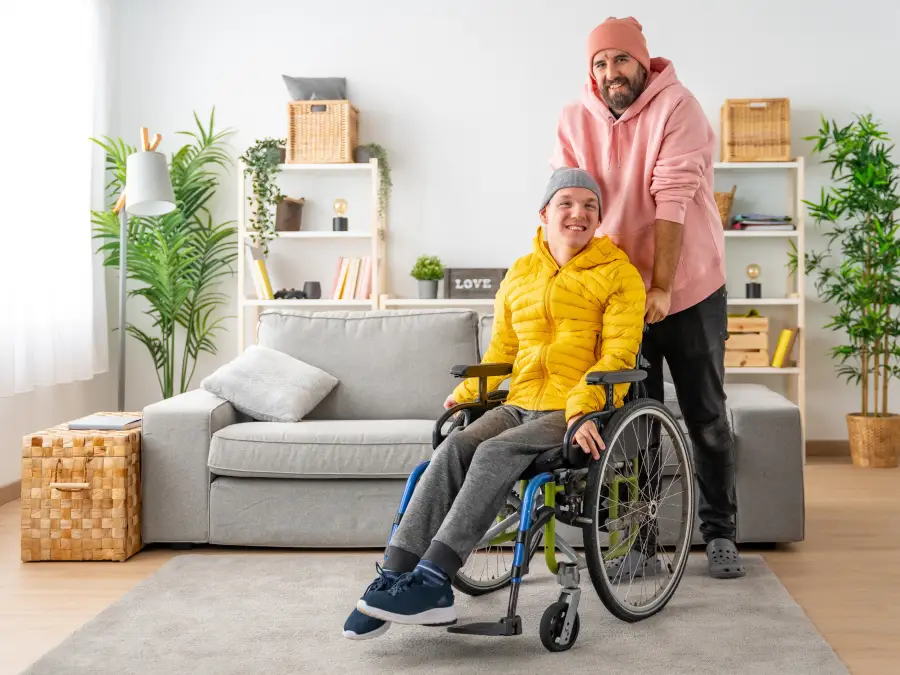 A portrait of a smiling caregiver standing behind a senior man in a wheelchair, both looking at the camera outdoors.