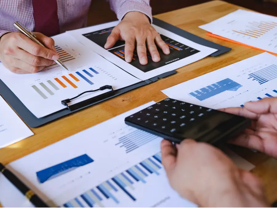 A team of business people sit at a desk, reviewing charts and data on a tablet and laptop during a meeting.