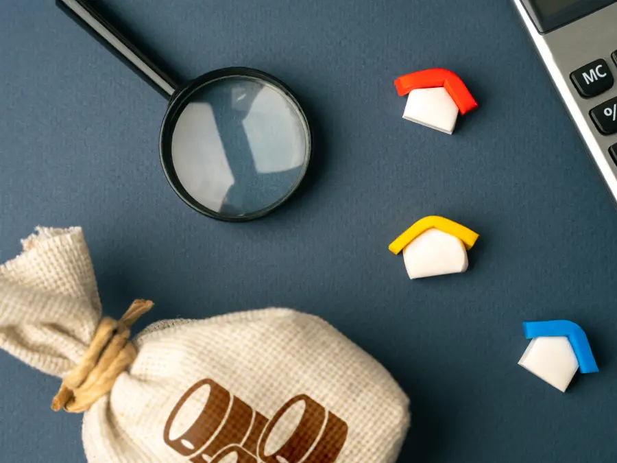 A flat layout of wooden toy houses, laptop, magnifying glass, and burlap of coins. 