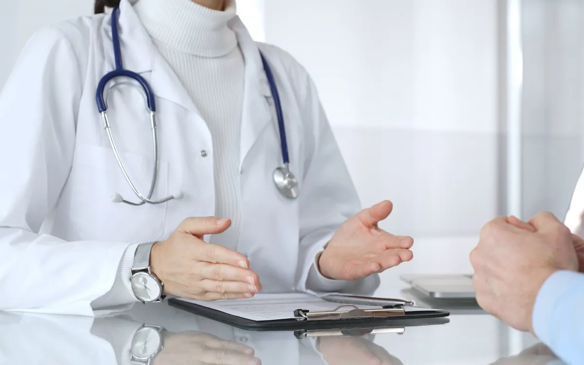 A close up shot of a doctor’s hand while wearing a stethoscope, explaining to a patient.