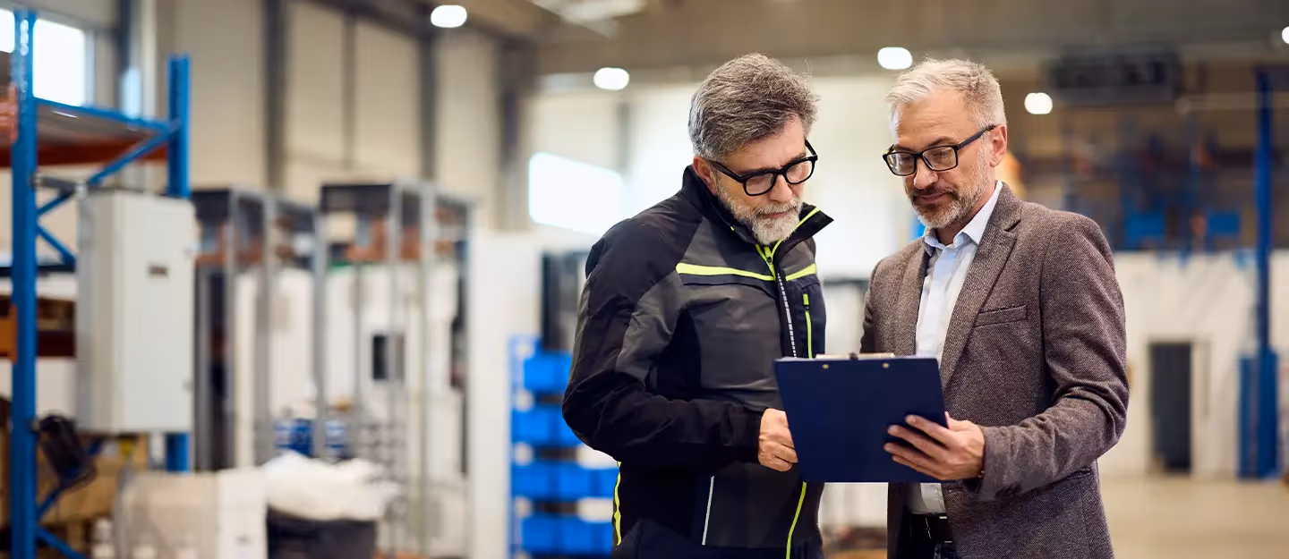 Two professionals standing in a modern manufacturing warehouse reviewing information on a clipboard beside industrial equipment and storage racks.