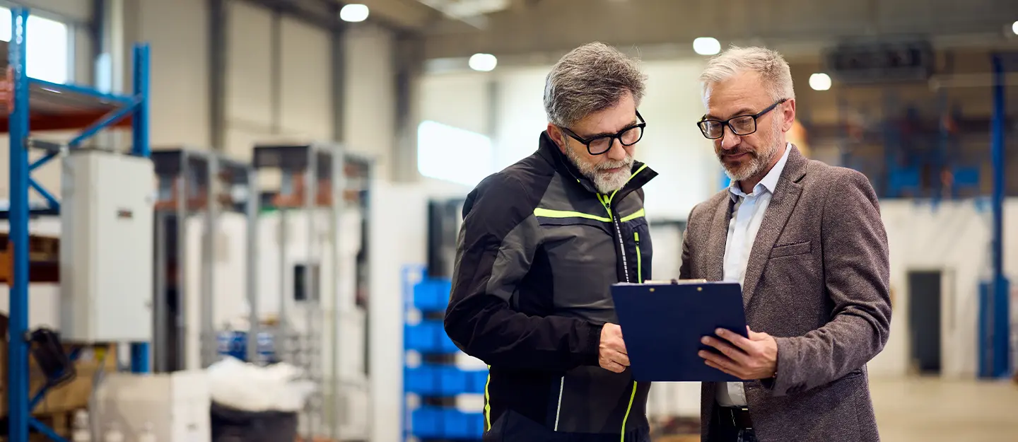 Two professionals standing in a modern manufacturing warehouse reviewing information on a clipboard beside industrial equipment and storage racks.