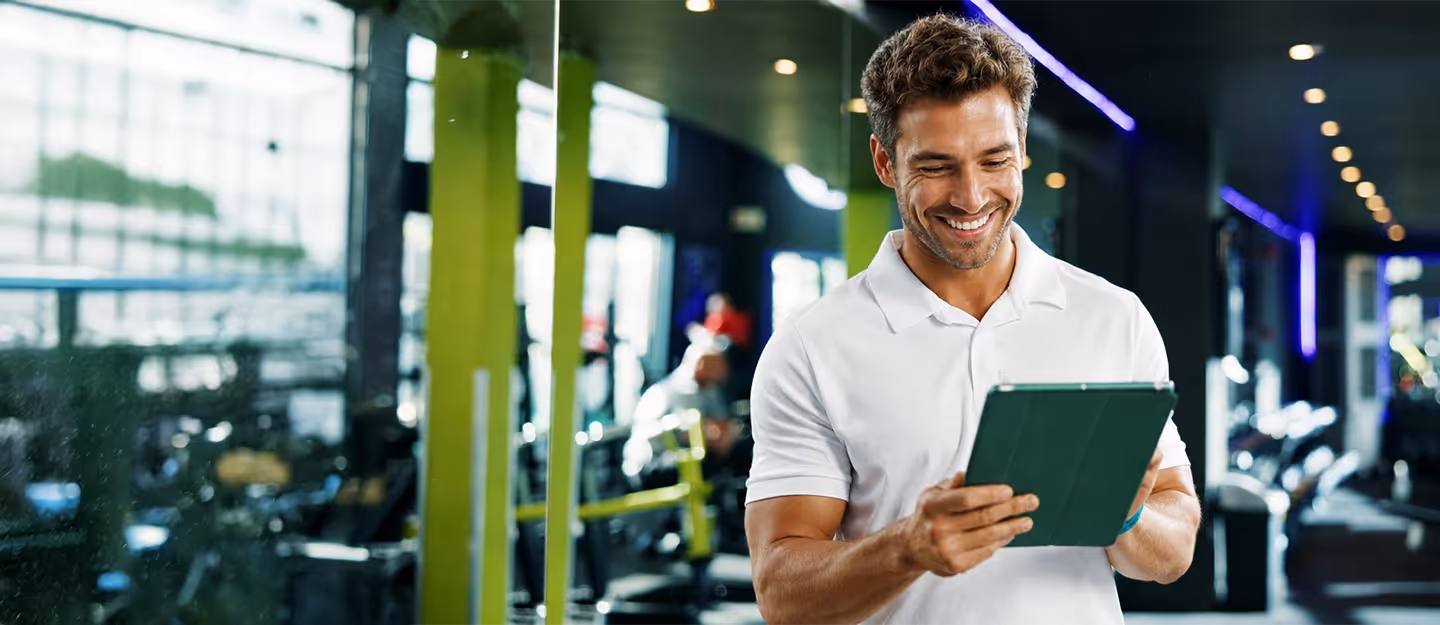 Smiling gym owner using a tablet inside a modern fitness center with exercise equipment in the background.