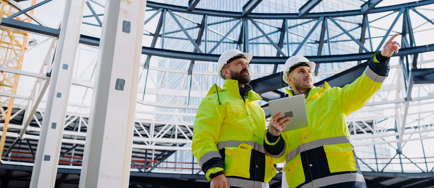 Two construction managers in high-visibility jackets and hard hats reviewing a building project on-site using a tablet inside a large steel-framed structure.