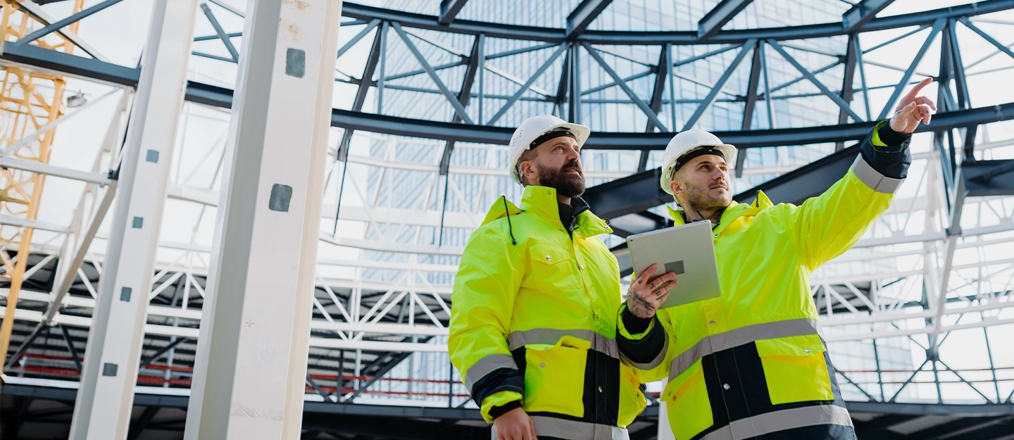 Two construction managers in high-visibility jackets and hard hats reviewing a building project on-site using a tablet inside a large steel-framed structure.