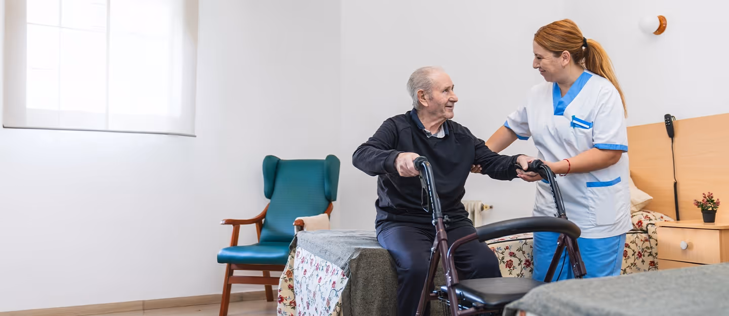 Aged care nurse assisting an elderly man with a walker beside a bed in a residential care facility room.