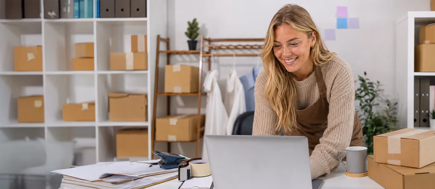 Ecommerce business owner working from a home office and preparing packaged orders while using a laptop.