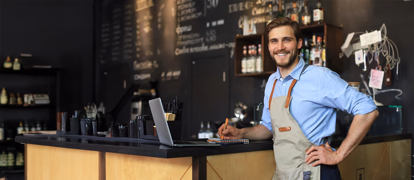 Bar owner standing behind the counter of a café while managing business operations with a laptop and notebook.