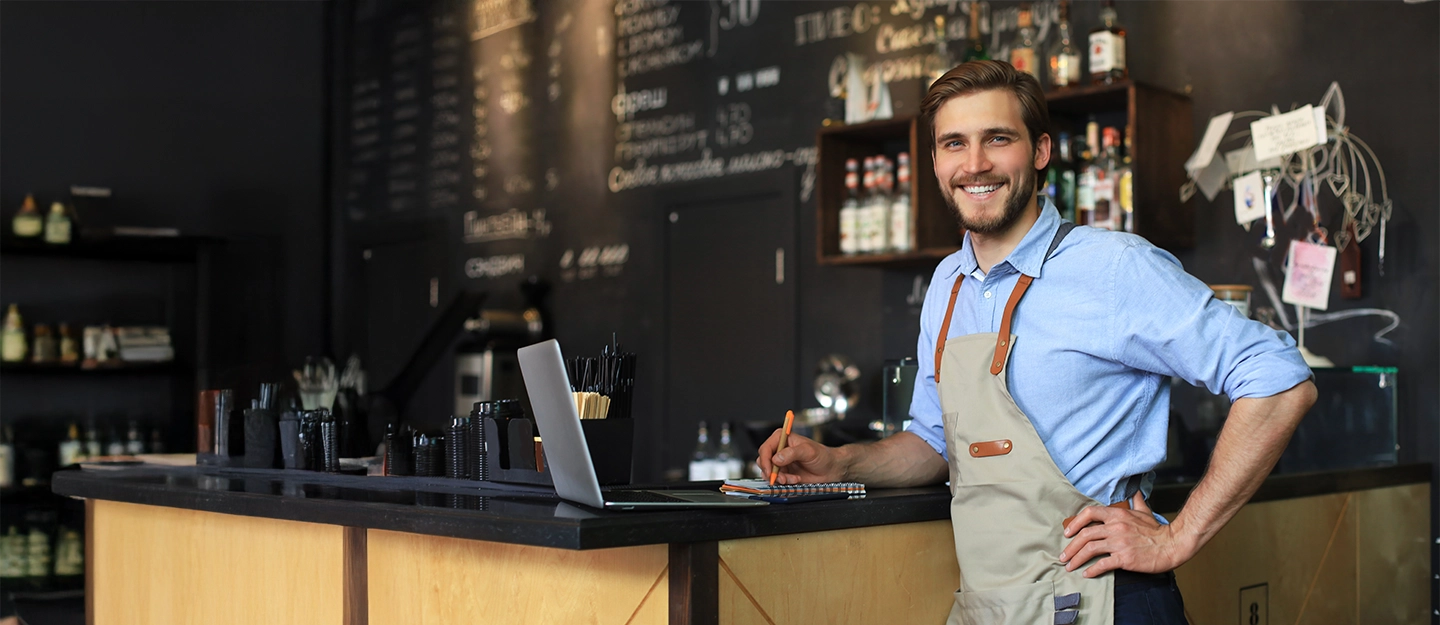 Bar owner standing behind the counter of a café while managing business operations with a laptop and notebook.