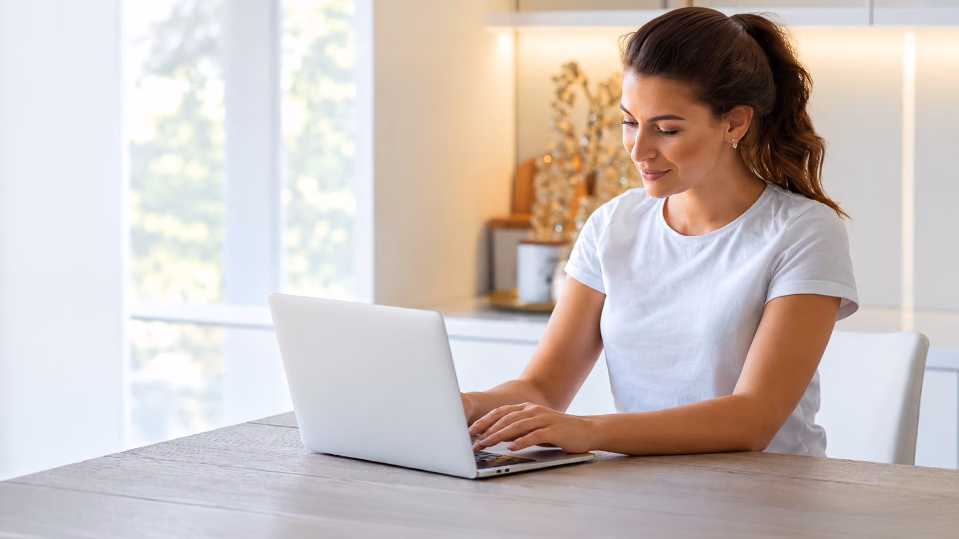 Smiling woman in a white t-shirt sitting at a table and typing on a silver laptop in a bright home office setting.