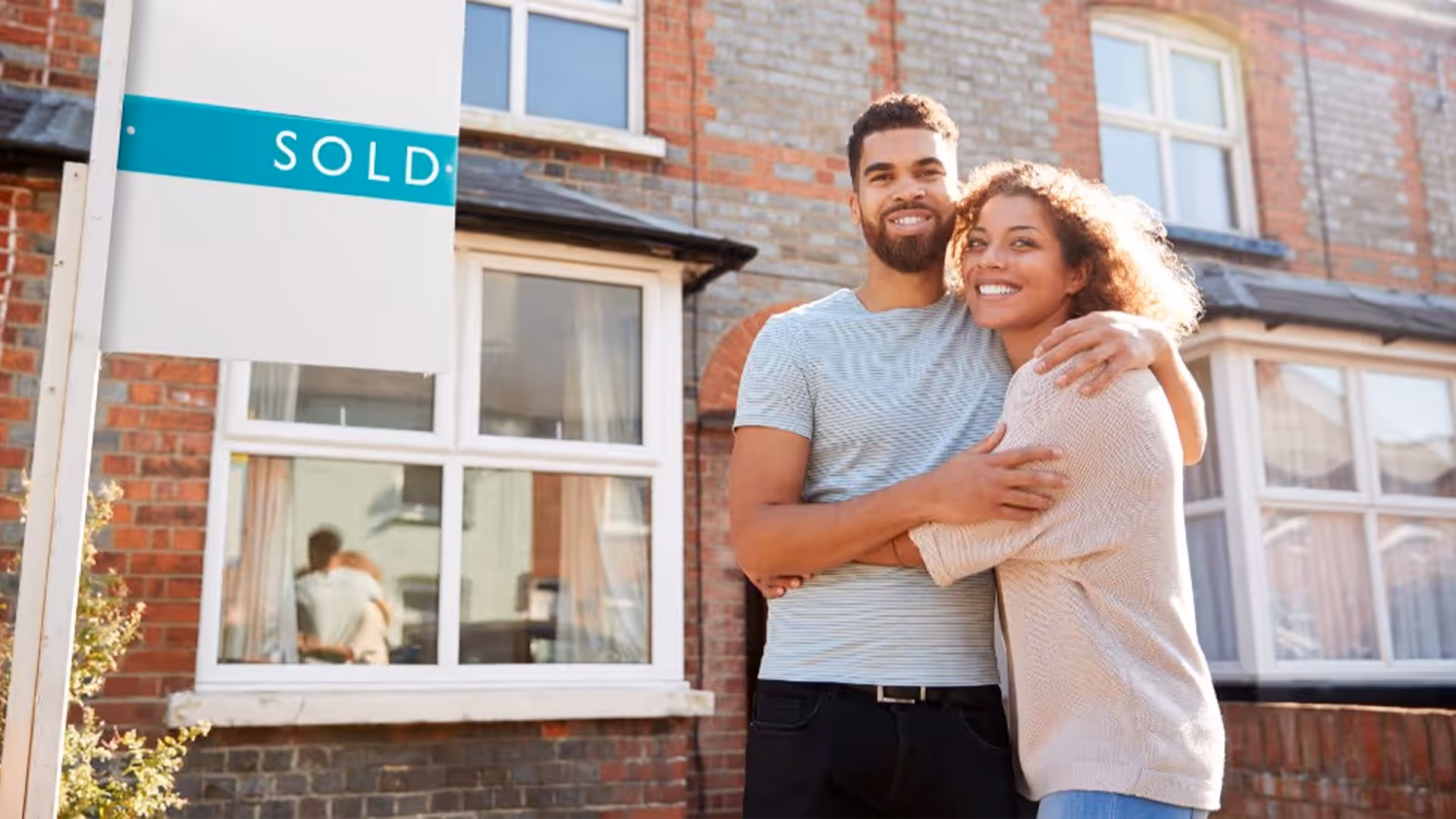 Smiling young couple embracing in front of a brick house with a white and teal real estate "SOLD" sign displayed.