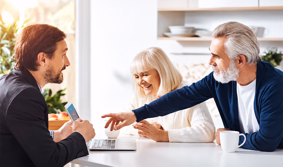 Senior couple meeting with an advisor at a table, with the man pointing towards a laptop screen in a bright room.
