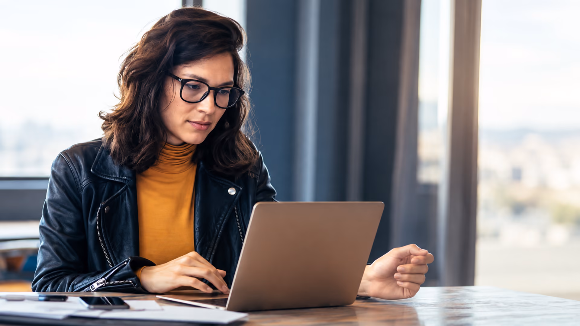 Woman wearing glasses, a yellow top and a leather jacket working on a laptop while sitting at a table in a modern space.