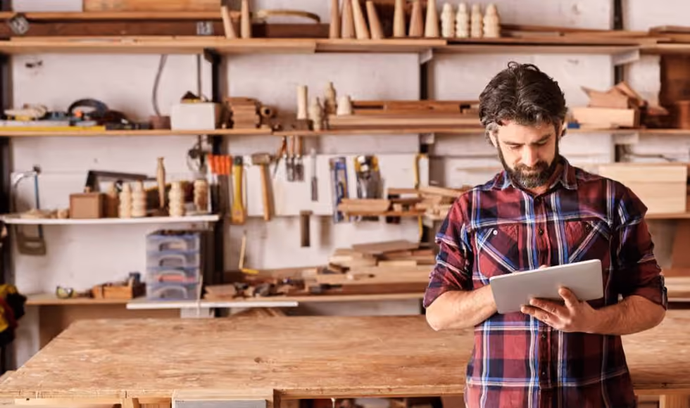 Male sole trader in a flannel shirt using a tablet computer inside a fully equipped woodworking workshop.