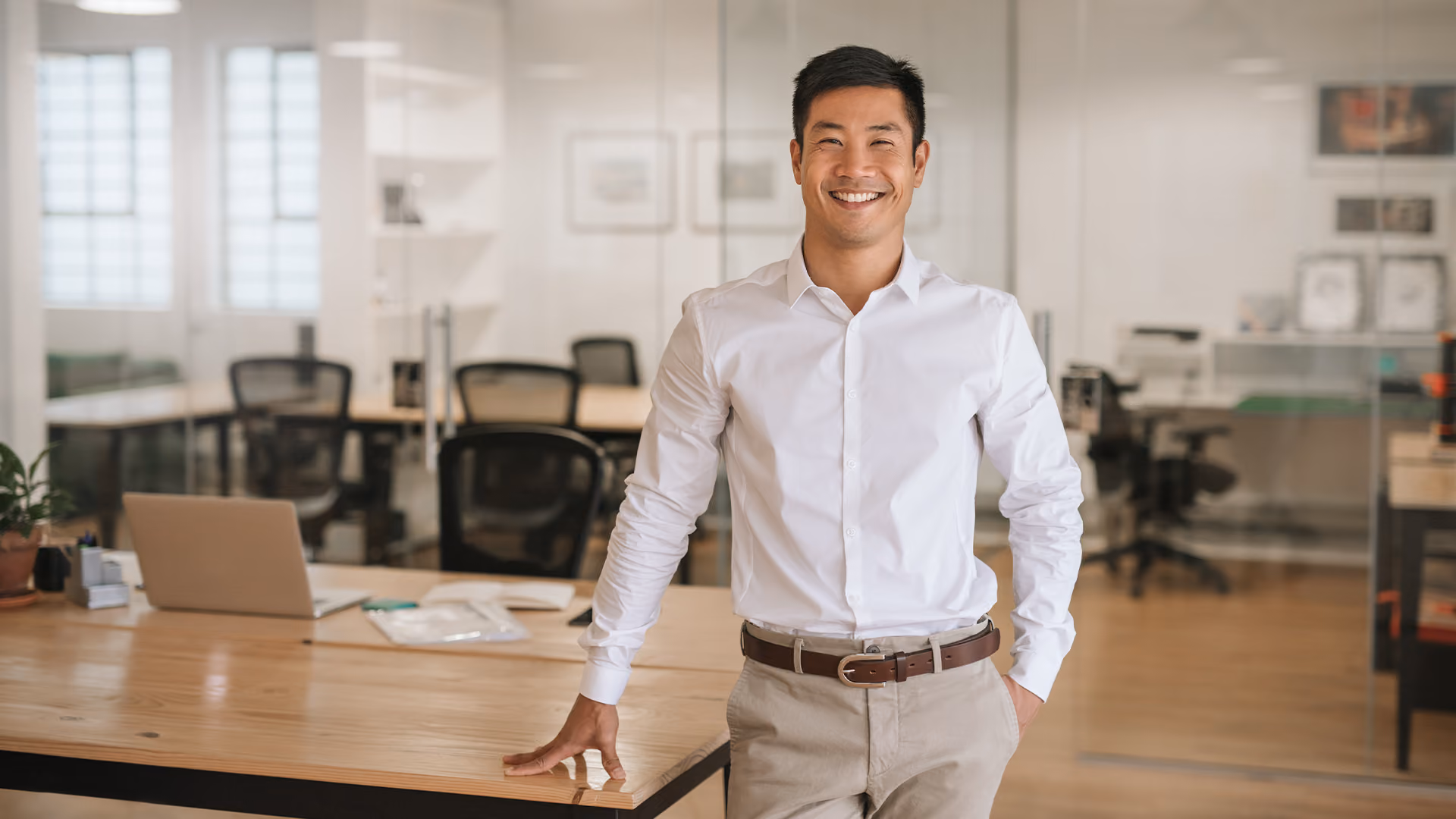 Smiling businessman leaning against a wooden desk in a bright, modern open-plan office space.
