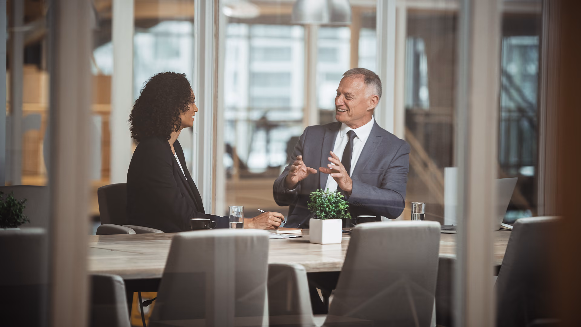 Senior businessman gesturing while conversing with a female colleague across a boardroom table in a modern office.