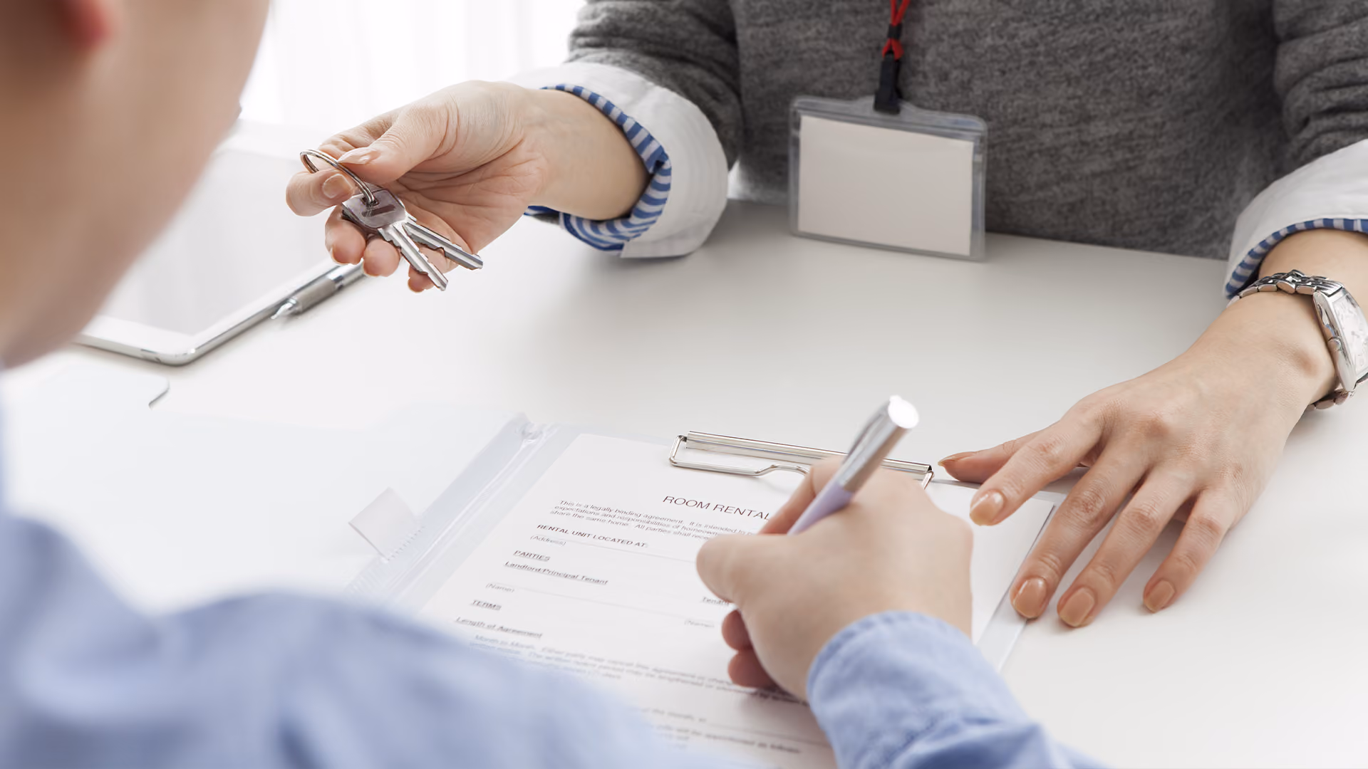 Real estate agent handing keys to a client signing a property rental agreement document over a desk.