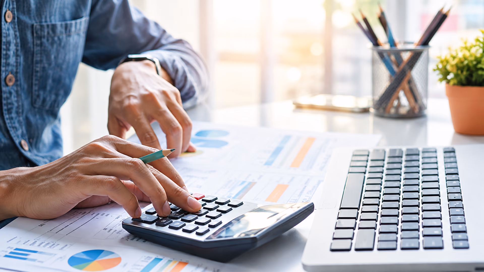 Person using a calculator with a pencil in hand while reviewing financial documents at a desk beside a computer keyboard.