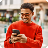 Smiling young man in a red sweater looking at his smartphone outdoors in an urban setting.