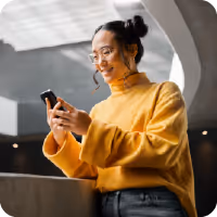 Smiling woman in a yellow sweater using a smartphone indoors with modern architecture in the background.