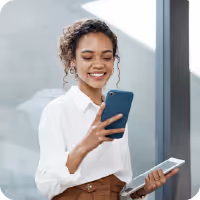 Smiling woman in white shirt holding a smartphone and a tablet near a window.