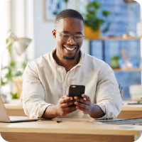 Smiling man in glasses and white shirt sitting at a desk using a smartphone.