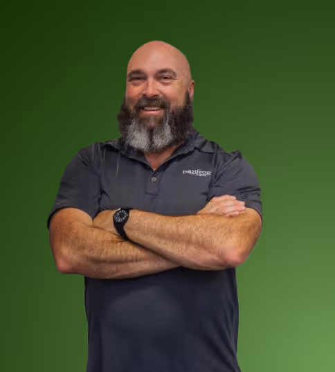 Smiling bald man with a gray beard wearing a dark polo shirt and a black wristwatch, standing with arms crossed against a green background.