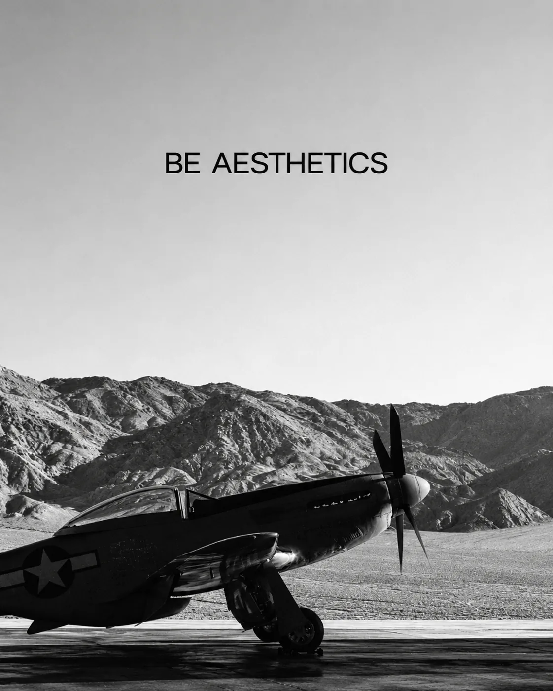 A black-and-white P-51 Mustang parked on a desert airfield below bare mountains, with the BE AESTHETICS wordmark in the sky