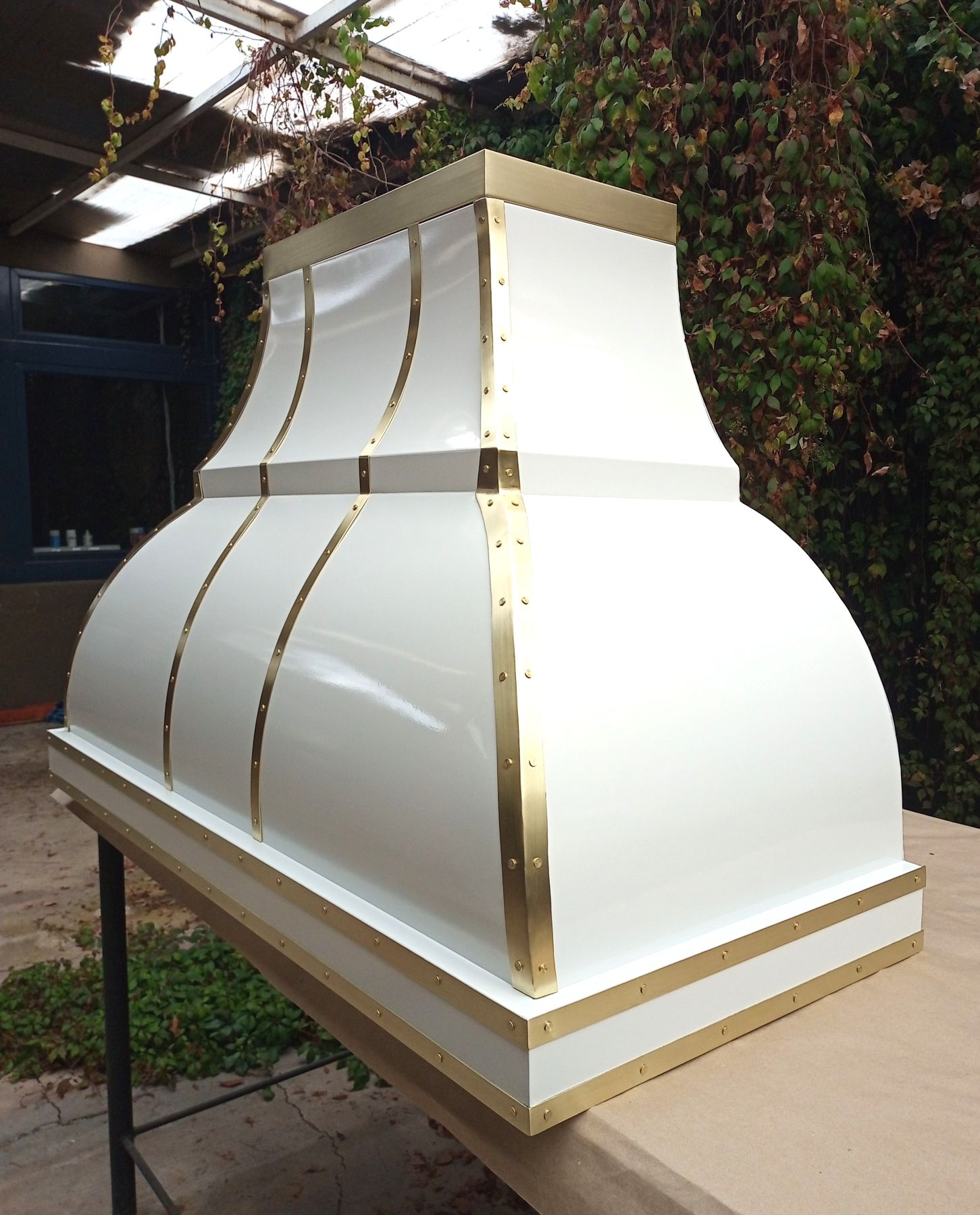 Custom white range hood with curved silhouette and brushed brass straps and rivets, Amoretti Brothers workshop, Mexico City