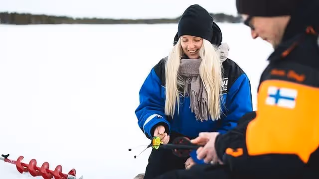 Smiling tourist in a blue winter jacket learning ice fishing techniques from a guide on a snowy frozen lake, holding a fishing rod, with an ice auger visible in the snow, in Rovaniemi, Lapland.