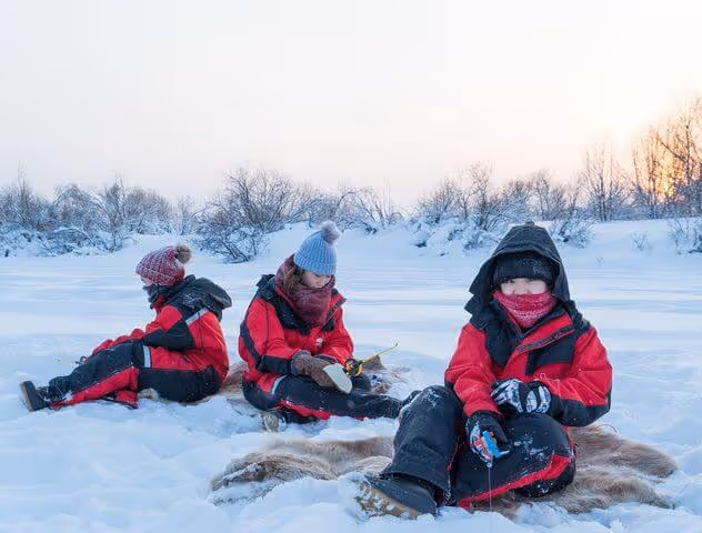 Children warmly dressed in red winter suits sitting on reindeer furs on a frozen, snowy lake, participating in an ice fishing activity during a winter day in Rovaniemi, Lapland.