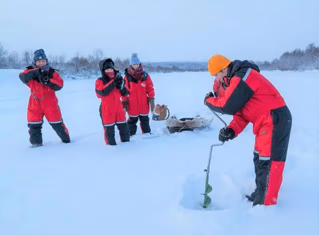 Combine a reindeer safari with in Lapland’s beautiful nature with an unforgettable ice fishing experience in Sierijärvi Lake. Group of people in red winter suits participating in an ice fishing activity on a vast, snow-covered frozen lake in Rovaniemi, Lapland, with one person drilling a hole in the ice.