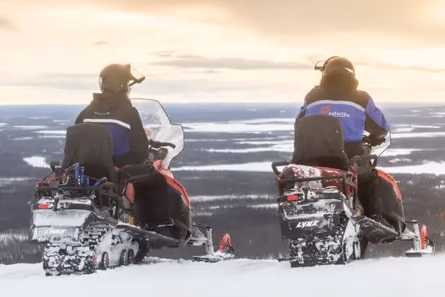 Two snowmobilers on their Lynx snowmobiles, looking out over a vast, snow-covered winter landscape with distant forest views, during a safari adventure in Rovaniemi, Lapland.