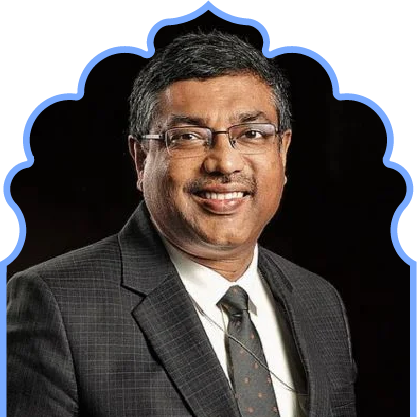 Smiling man with glasses wearing a dark checkered suit, white shirt, and patterned tie against a black background.