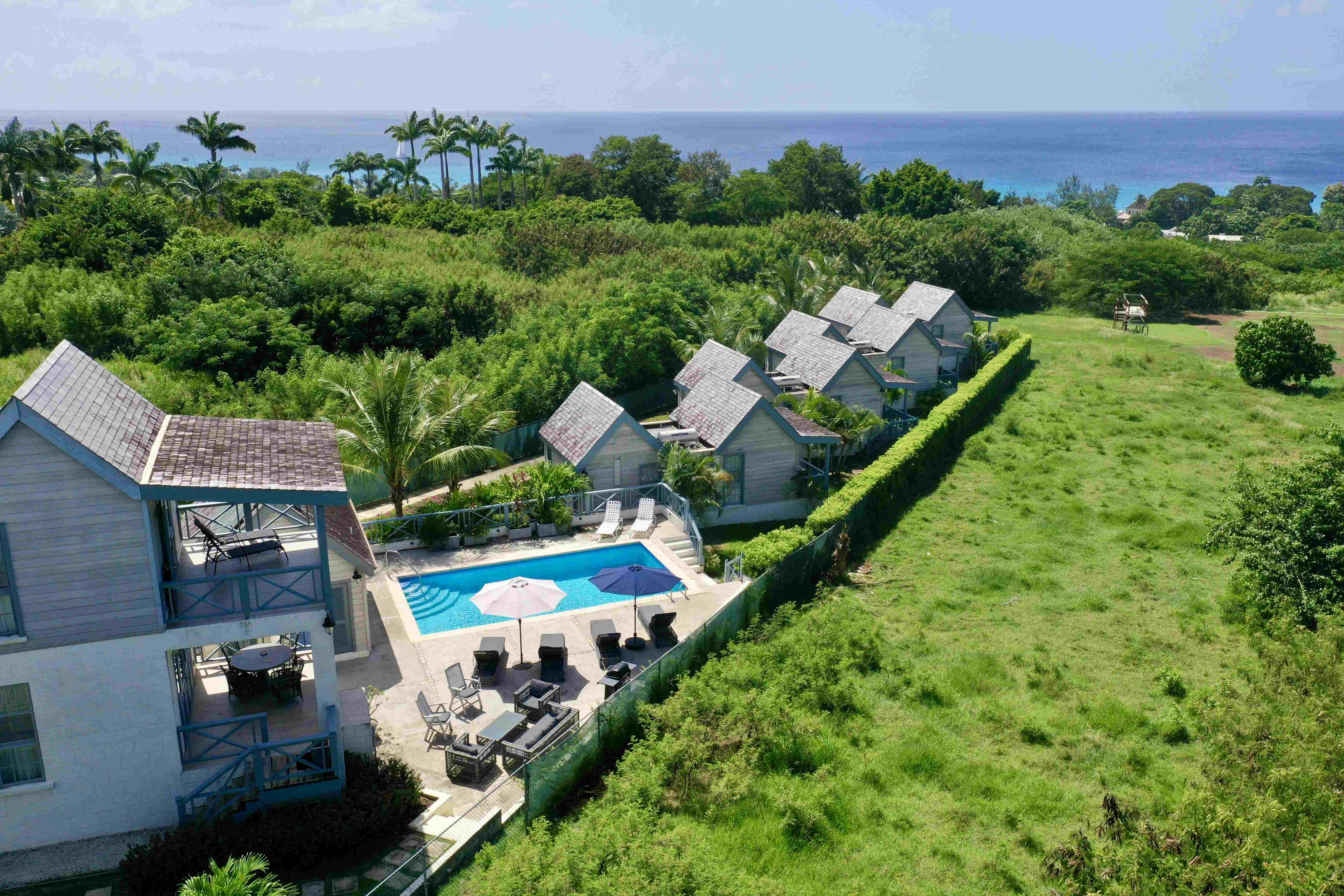 Aerial view of a resort with small cottages, a swimming pool with umbrellas and lounge chairs, surrounded by green vegetation and overlooking the ocean.