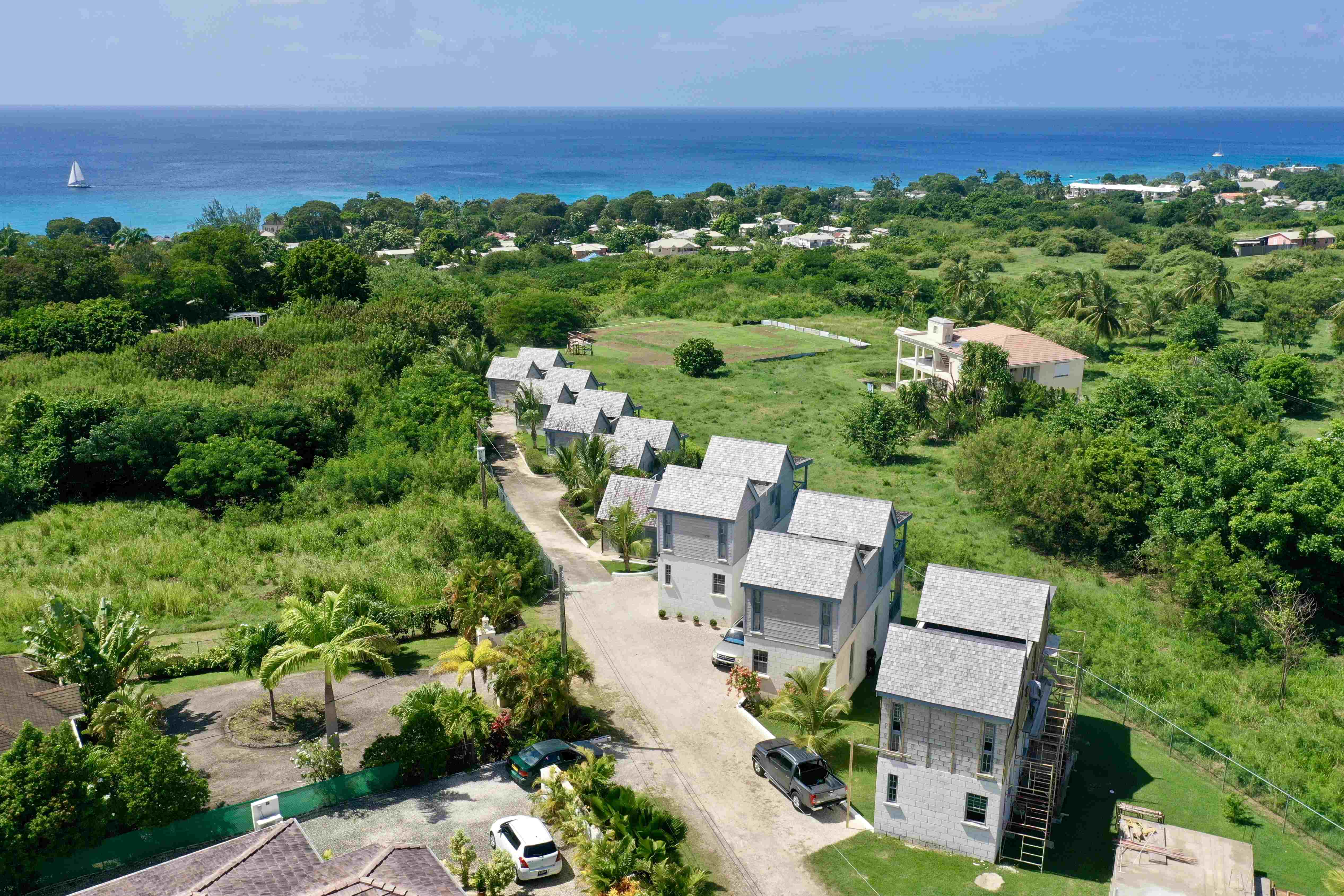 Aerial view of a coastal residential area with several gray-roofed houses, lush green vegetation, and the blue ocean in the background.