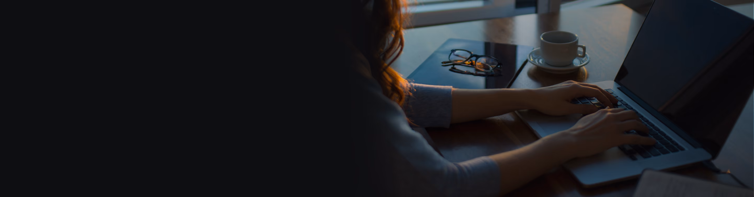 Person typing on laptop with eyeglasses and cup on table in soft natural light by window.
