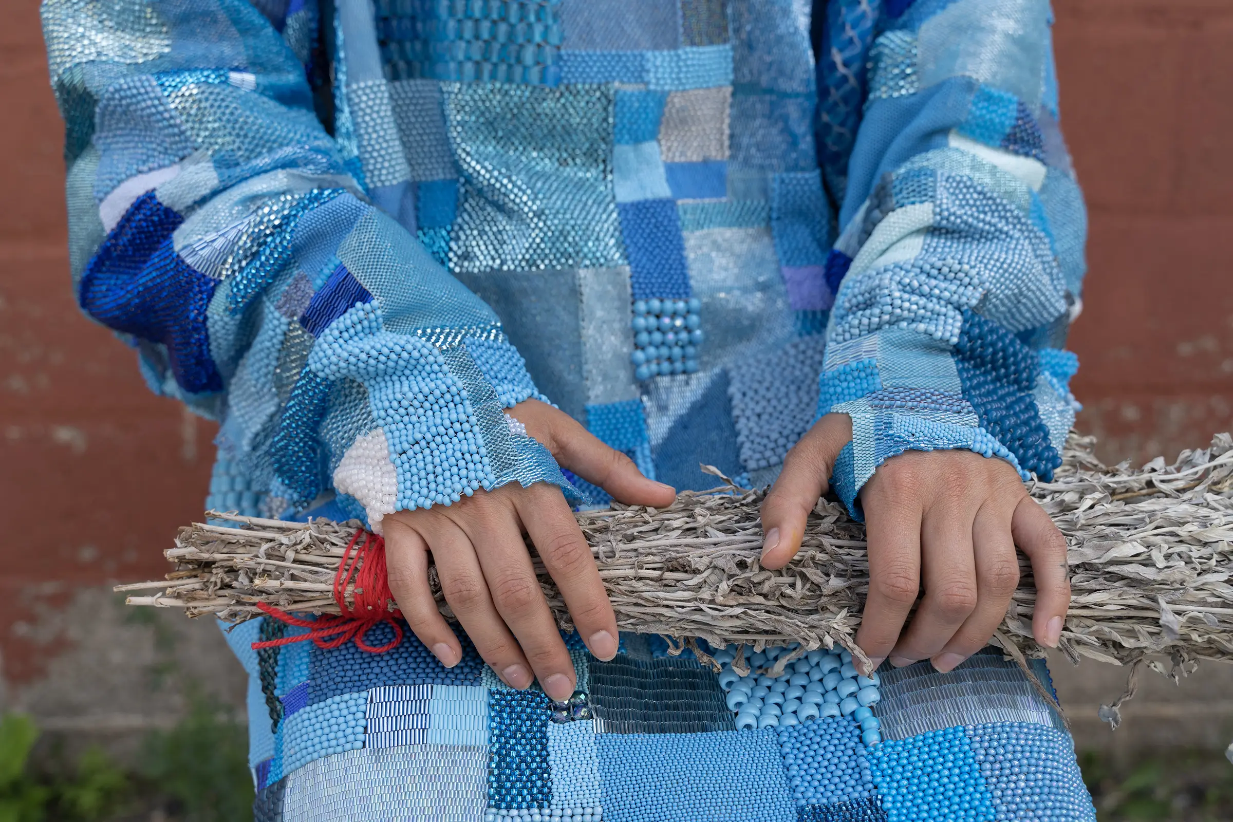 Close-up of someone wearing Maggie Thompson’s blue beaded patchwork Hospital Gown, their hands holding a bundled object while the intricate blue textiles and beadwork are shown in detail.