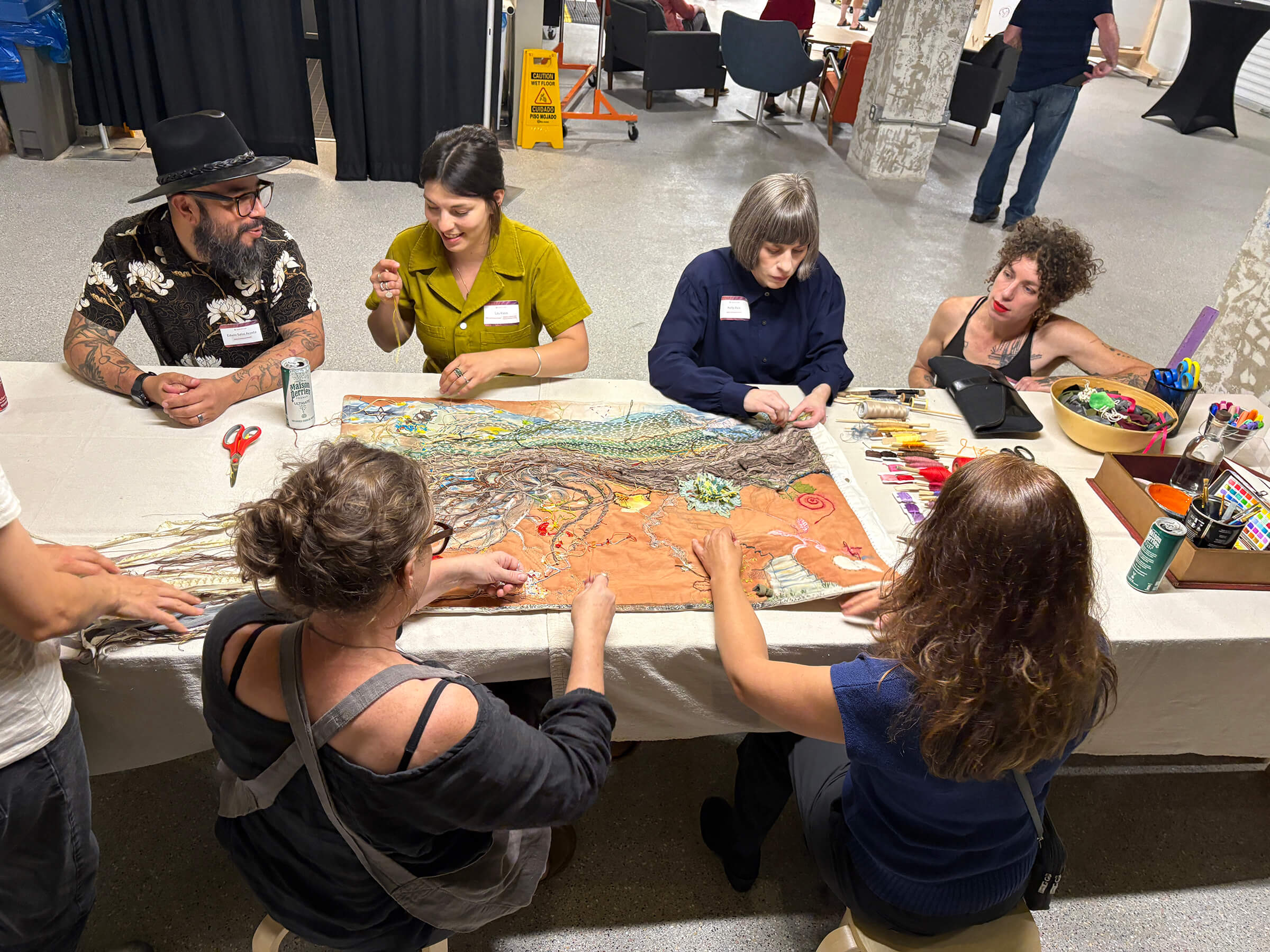 A group of people stand and listen to a person giving a demonstration at a table in a well-lit indoor space with string lights.