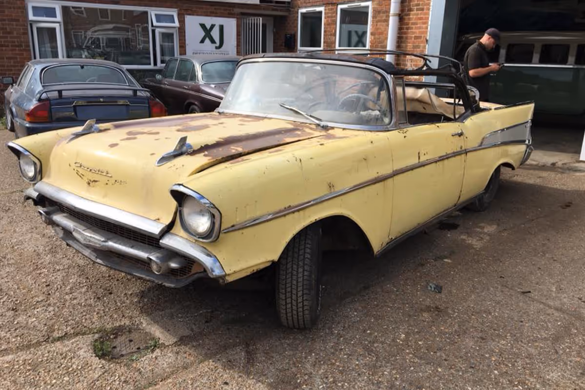 A rusty yellow vintage convertible car parked in front of a garage, with a person nearby.