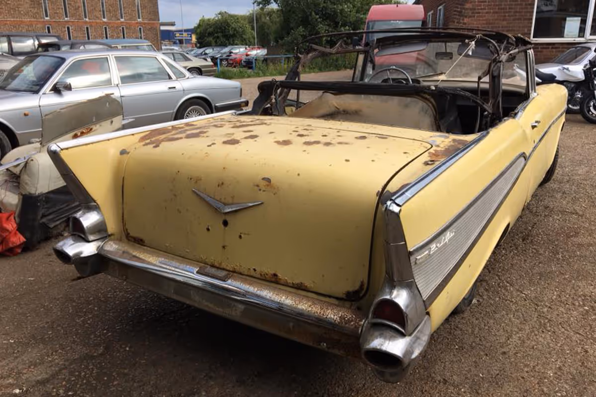 A rusty, yellow classic convertible car with a damaged roof is parked in a lot.
